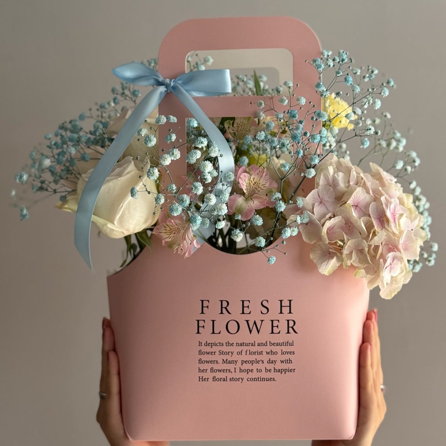 Pink box with flowers and a blue bow held by hands against a plain background
