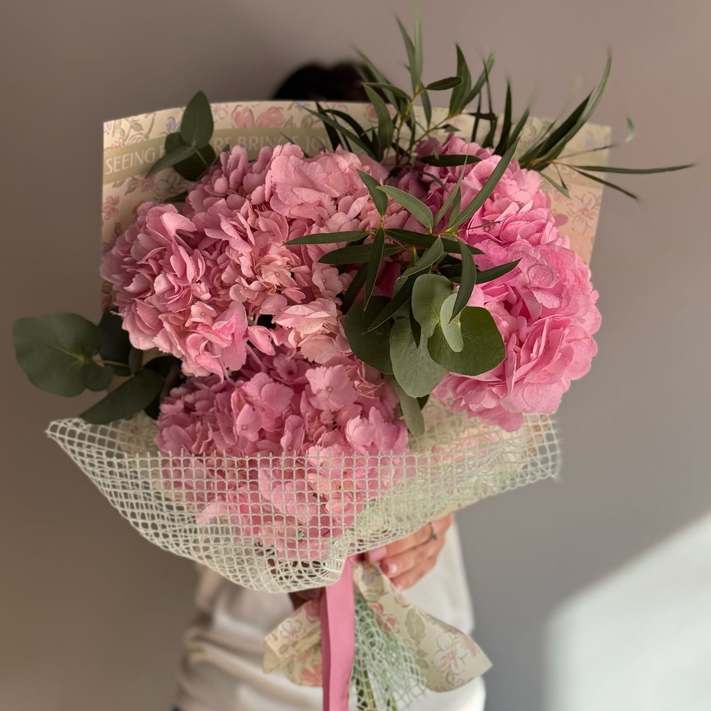 Bouquet of pink flowers wrapped in netting with a pink ribbon, held by a person against a neutral background.