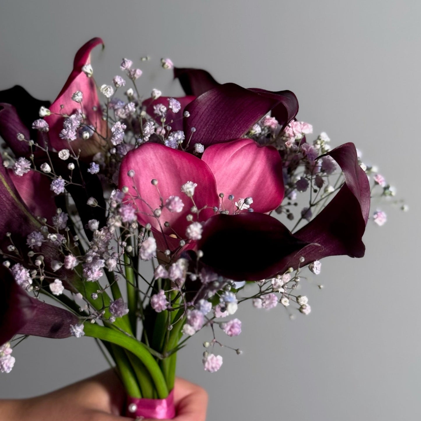 Bouquet of purple calla lilies with baby's breath held by a hand against a gray background