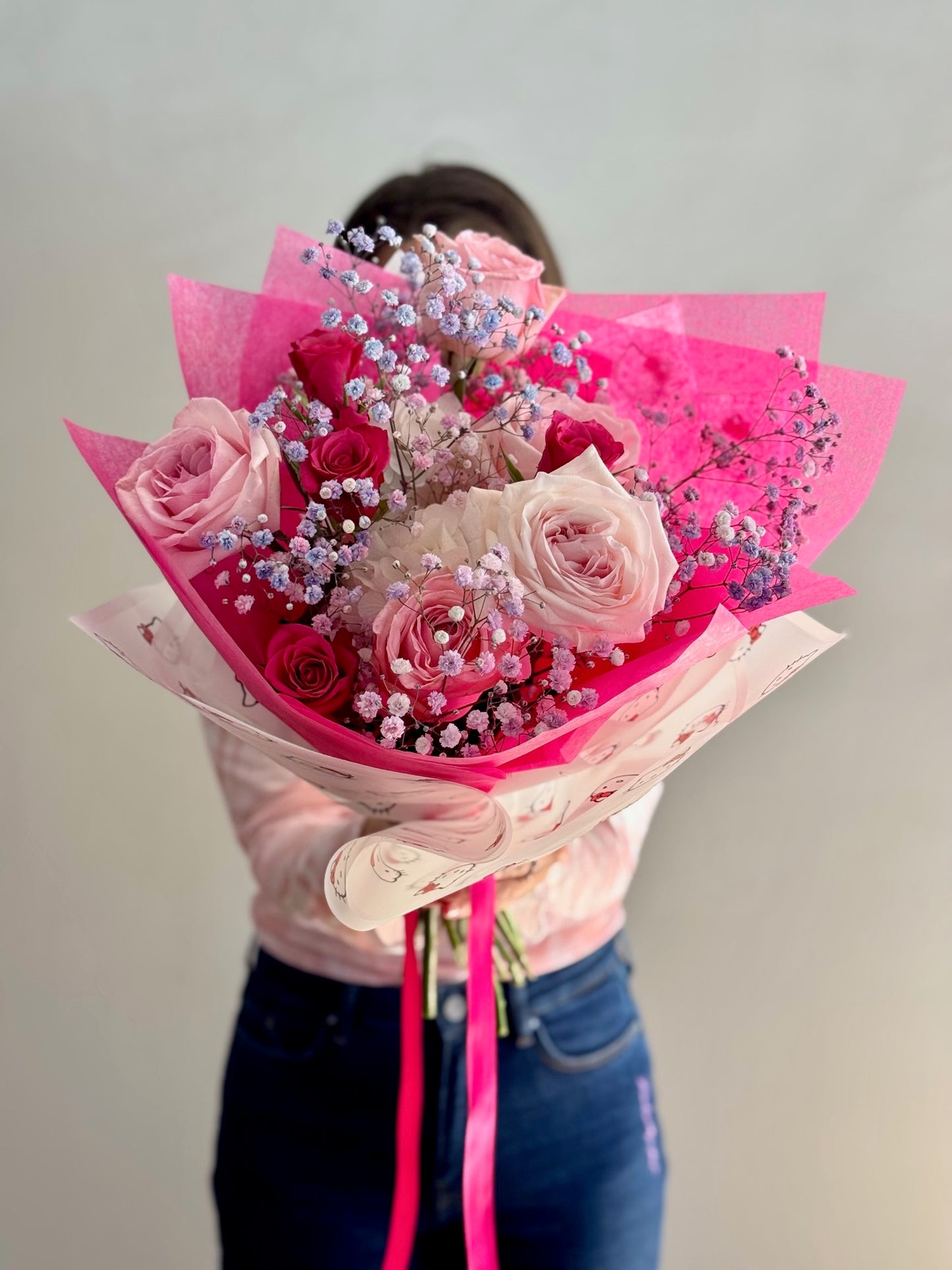 Person holding a bouquet of pink roses and flowers wrapped in pink paper against a plain background