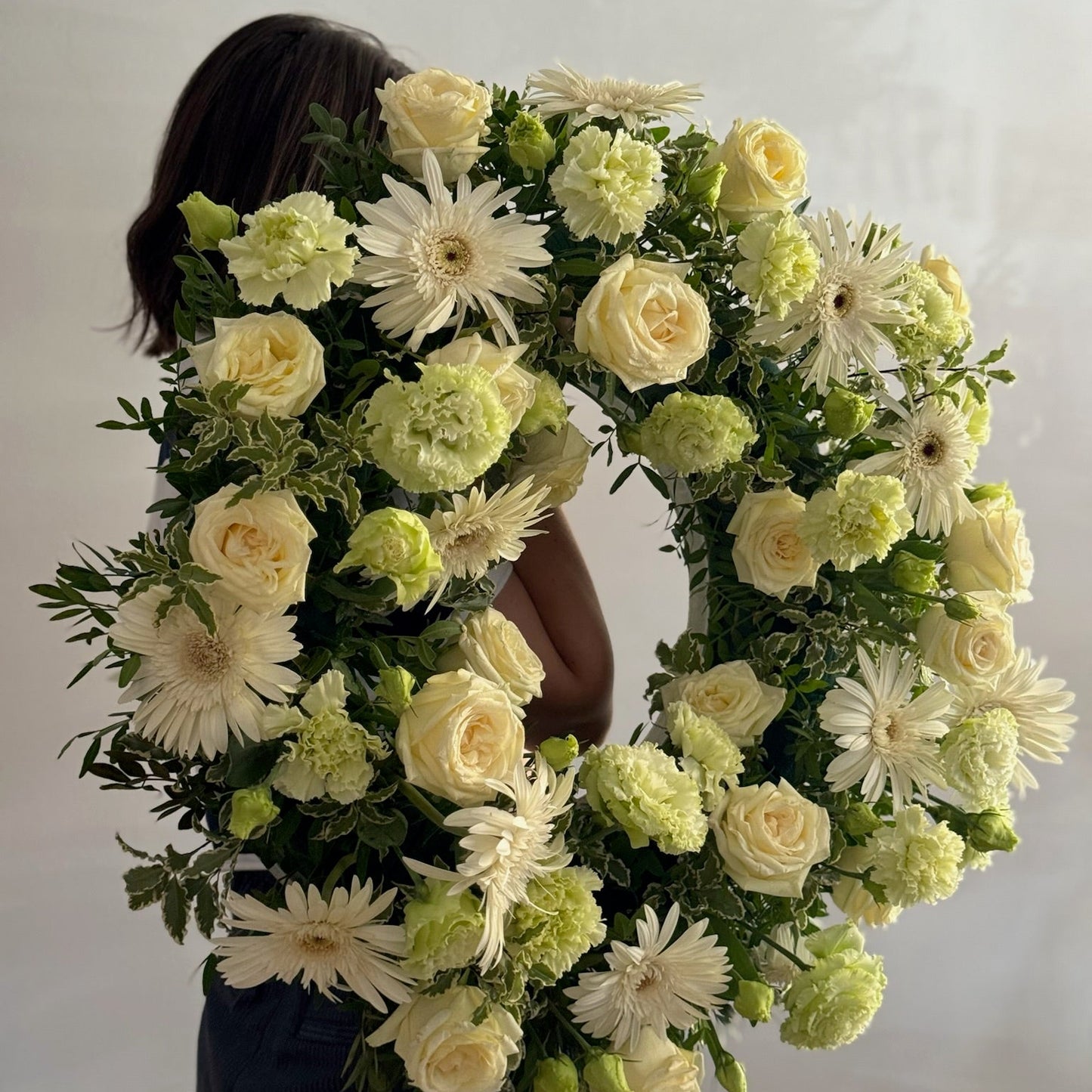 Person holding a large floral wreath made of white and green flowers against a plain background