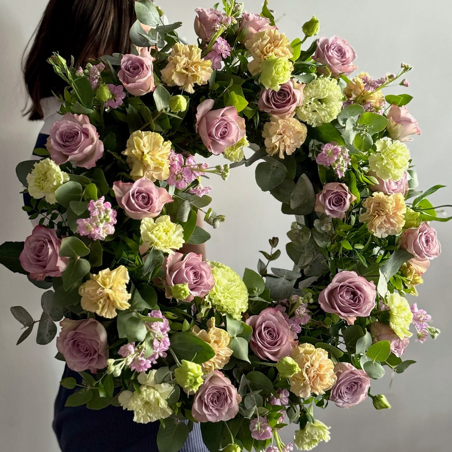 Person holding a large floral wreath with pink, yellow, and green flowers against a plain background
