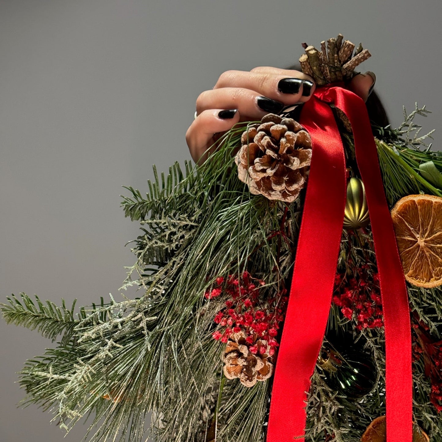 Decorative Christmas arrangement with greenery, pinecones, red ribbon, and dried oranges on a gray background.
