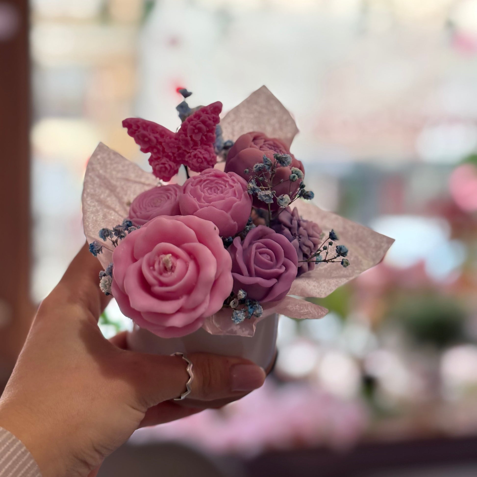 Hand holding a small bouquet with pink and purple flowers and a butterfly against a blurred background.
