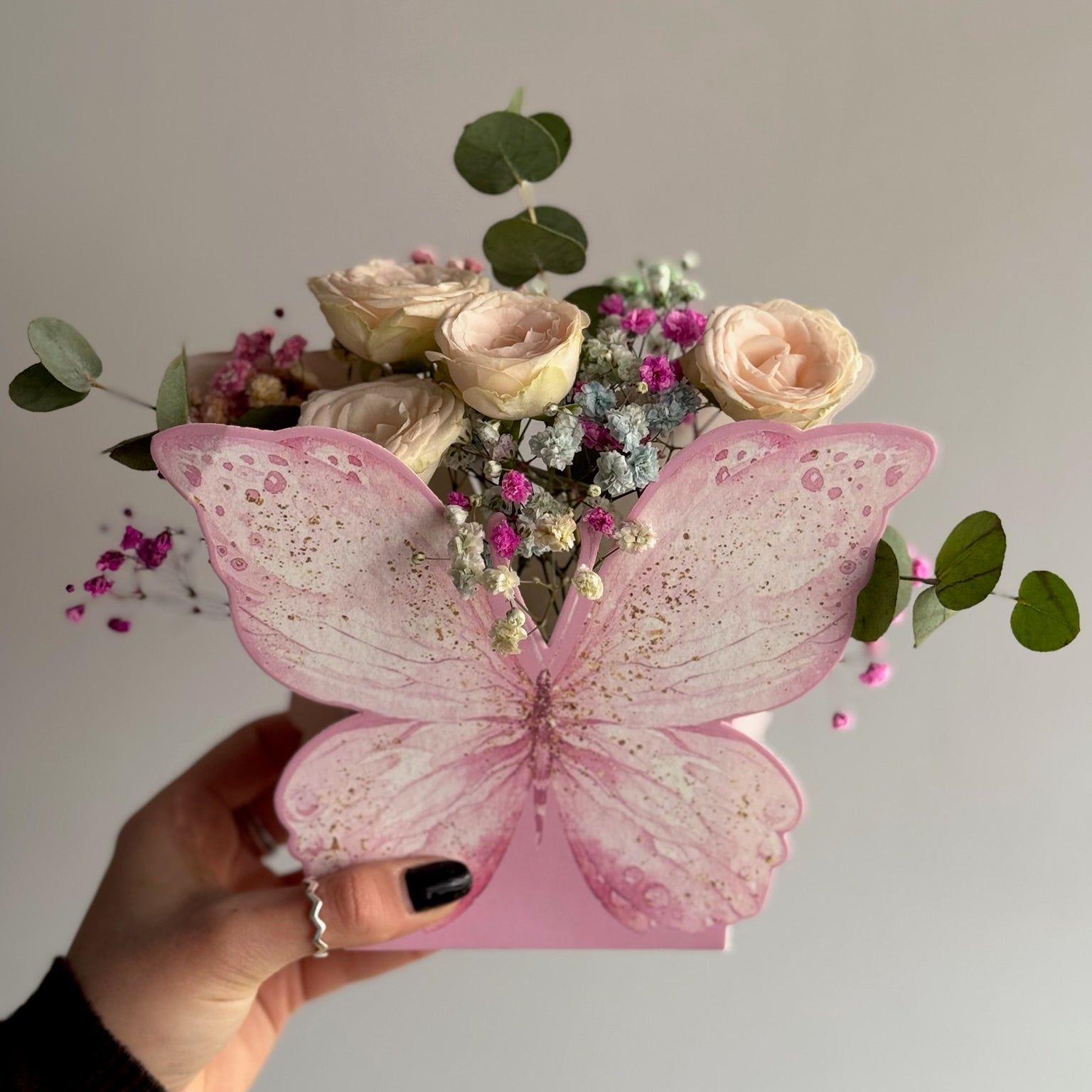 Bouquet of flowers in a pink butterfly-shaped box held by a hand against a neutral background