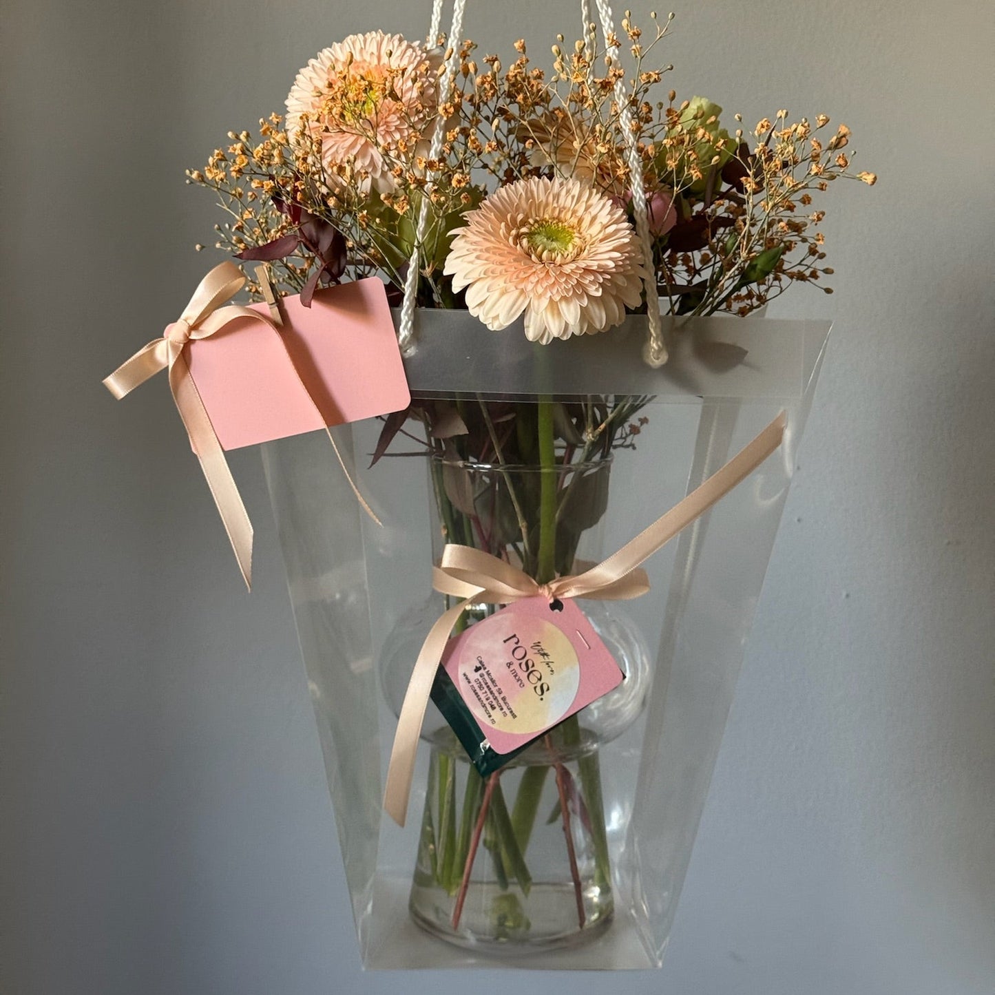 Bouquet of flowers with a pink gift box and tag, held by a hand against a neutral background.