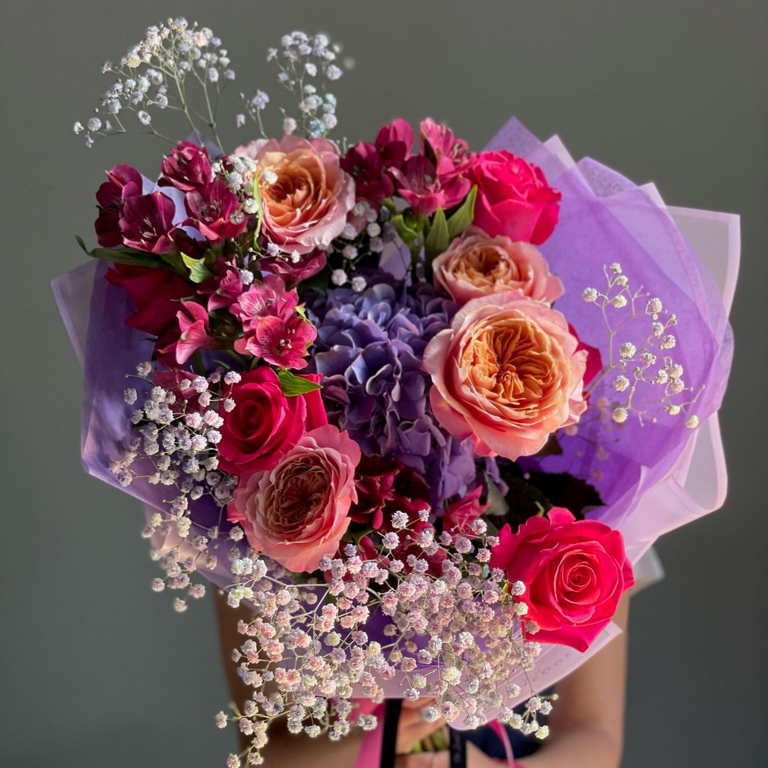 Bouquet of colorful flowers with a purple wrap held by a person against a gray background