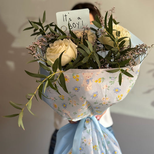Bouquet of flowers with a baby announcement card held by a person against a neutral background