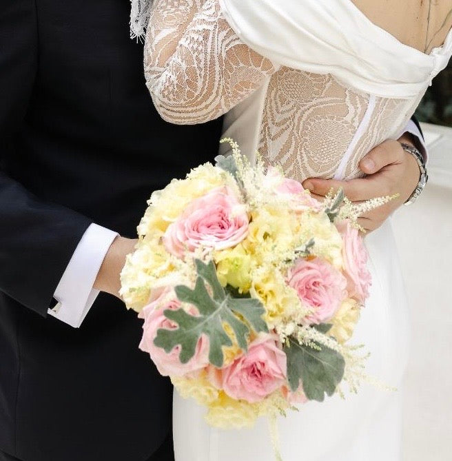 Bride holding a bouquet of pink and yellow flowers with a groom in the background.