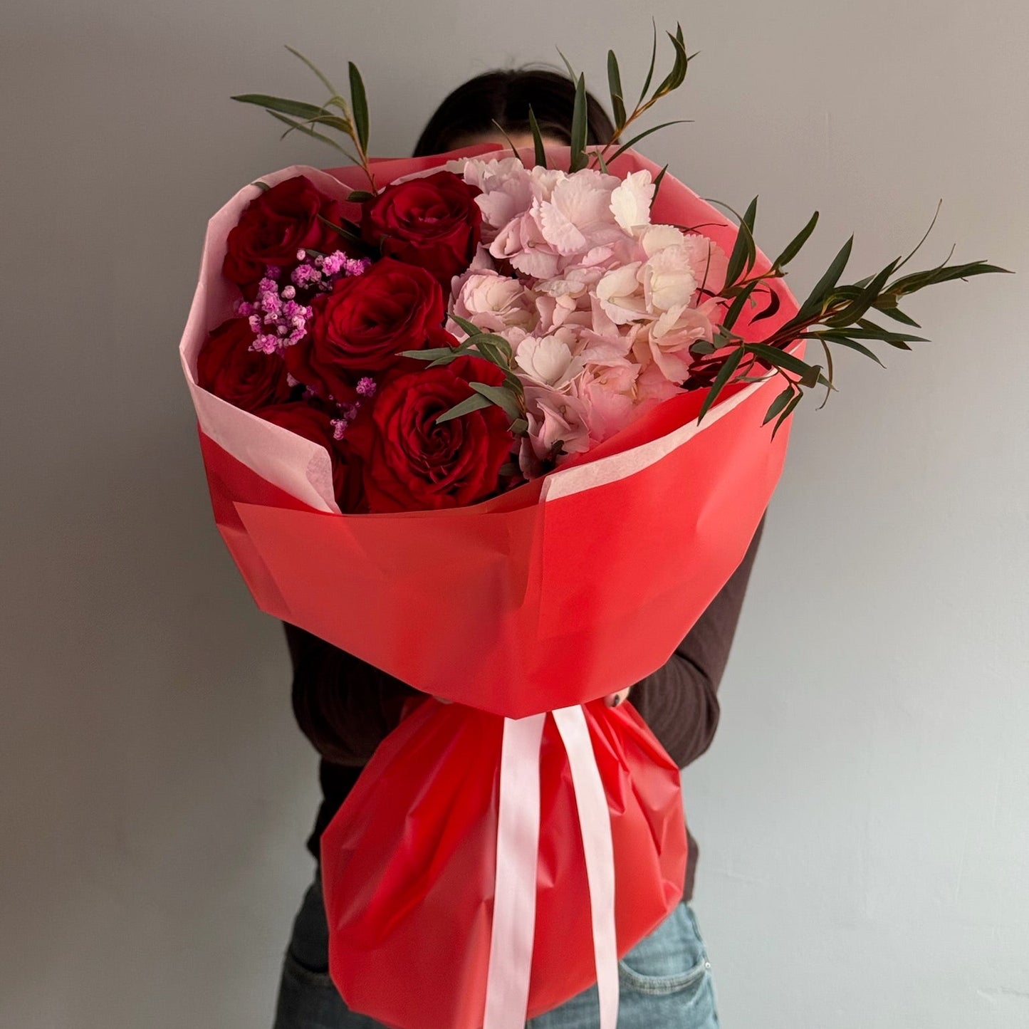 Person holding a bouquet of red and pink flowers wrapped in red paper against a plain background