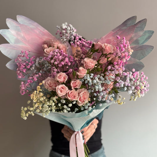 Bouquet of pink and white flowers with sheer pink and blue ribbons held by a person against a neutral background.