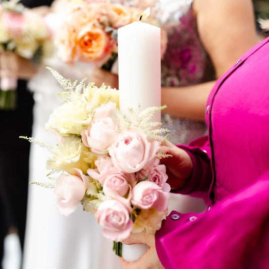 Person holding a bouquet with pink flowers and a candle, blurred background of people in formal attire.