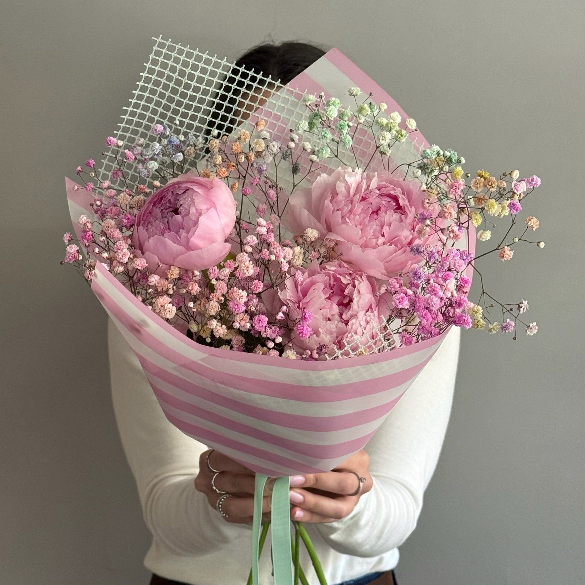 Pink peonies bouquet in cute pink wrapping paper held by person on grey background 