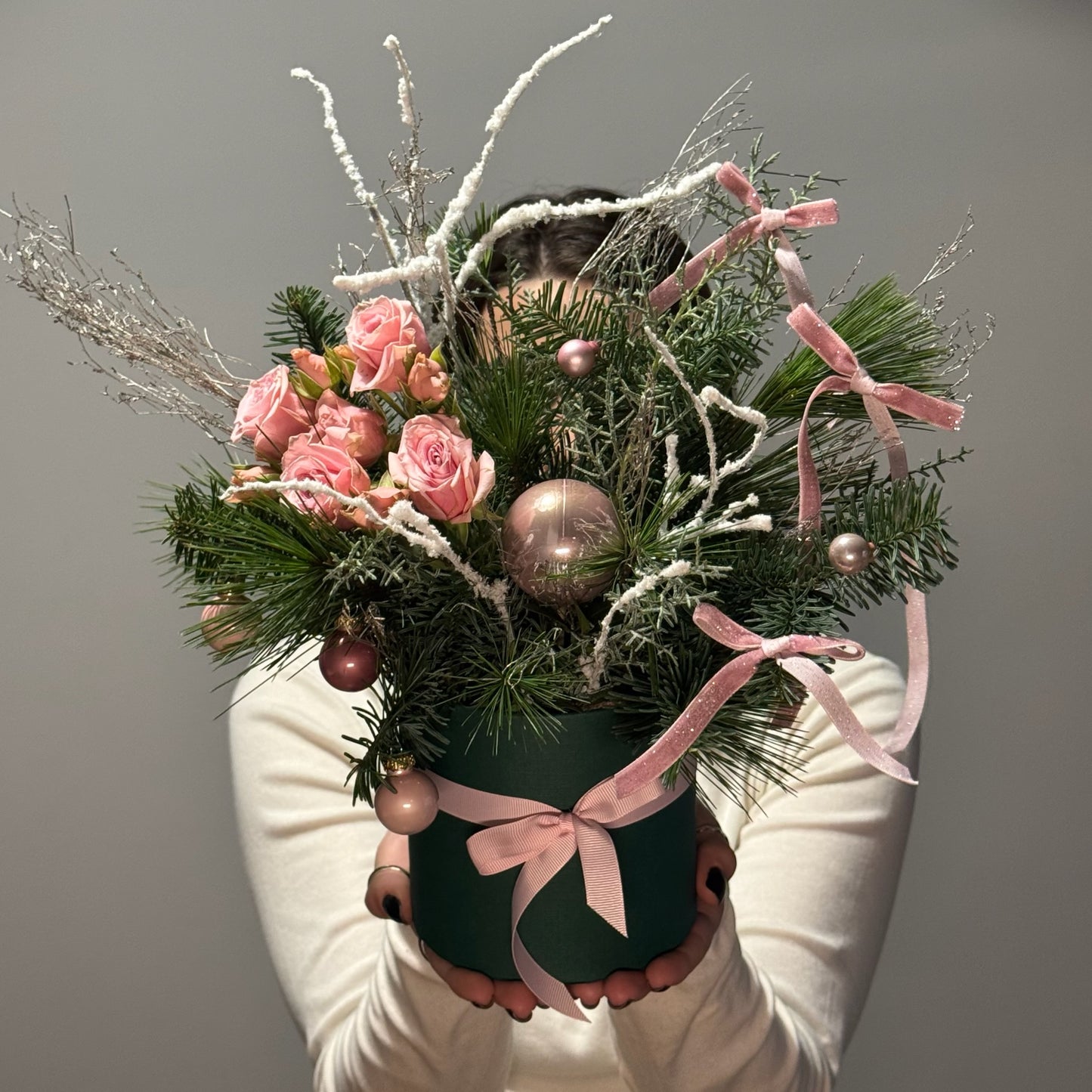 Floral arrangement with pink roses, greenery, and ornaments held by a person wearing white gloves against a gray background.