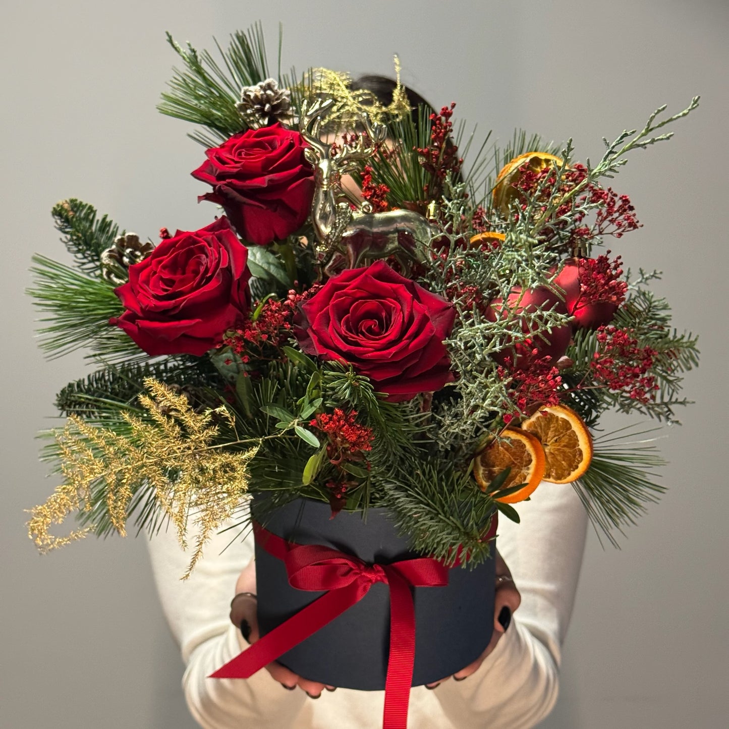 Bouquet of red roses and greenery with a black box and red ribbon held by a person against a gray background