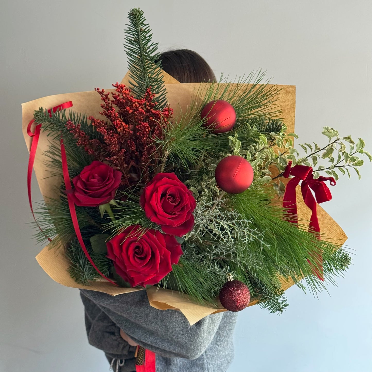 Bouquet of red roses and greenery with red ornaments and ribbons, held by a person.