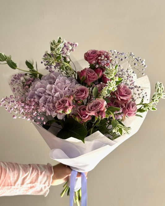 Bouquet of purple and pink flowers wrapped in white paper, held by a person wearing gloves.