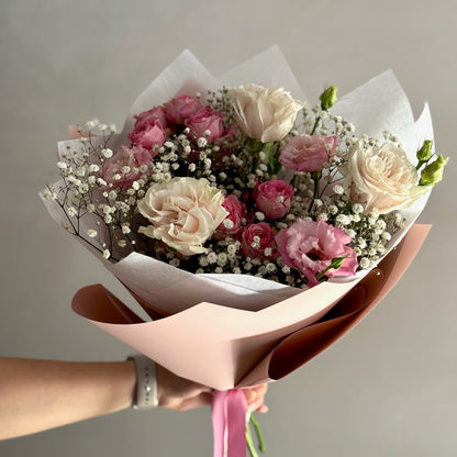 Bouquet of pink and white flowers wrapped in brown paper with a pink ribbon held by a person against a plain background.