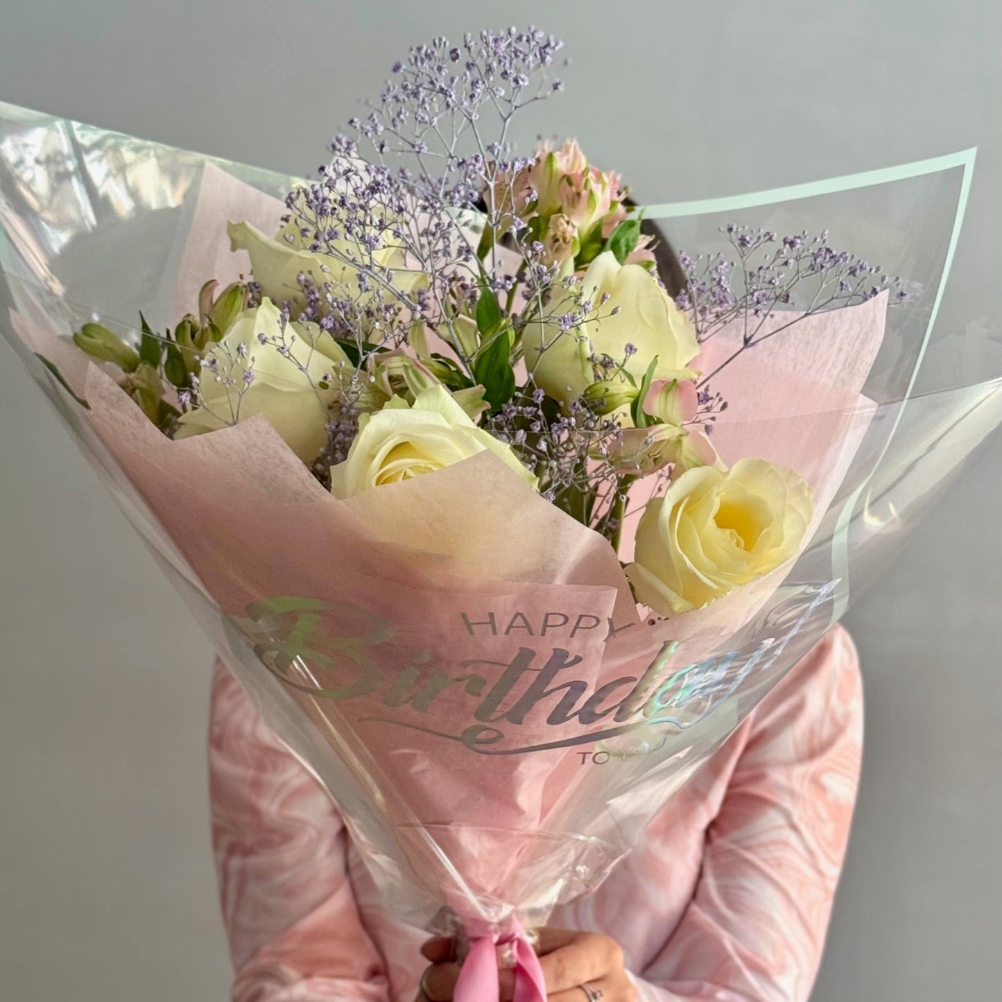 Bouquet of flowers wrapped in pink paper with 'Happy Birthday' text, held by a person wearing a pink jacket.
