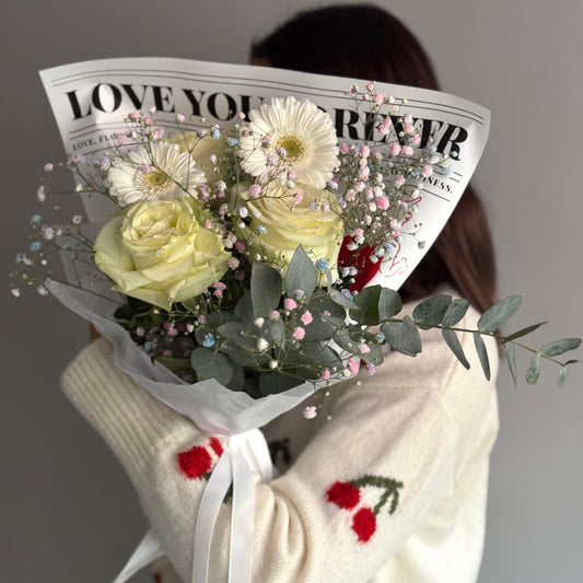 Person holding a bouquet of flowers wrapped in newspaper with 'LOVE YOU FOREVER' printed on it, wearing a white sweater with red patterns.
