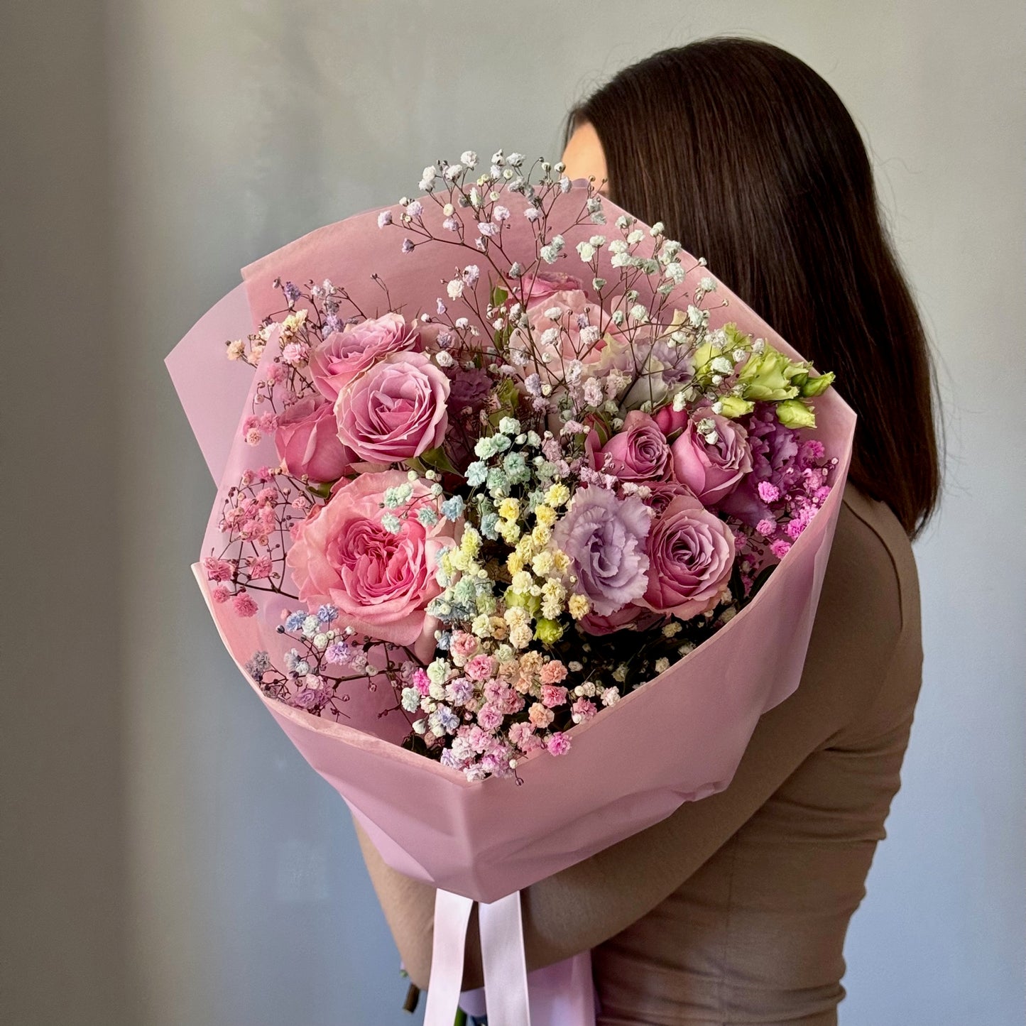 Person holding a bouquet of pink and purple flowers wrapped in pink paper against a neutral background