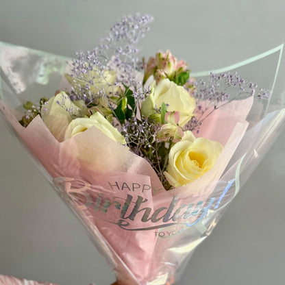 Bouquet of yellow roses wrapped in pink paper with 'Happy Birthday' text, held by a person wearing gloves.