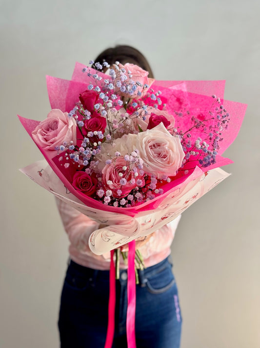 Person holding a bouquet of pink roses and flowers wrapped in pink paper against a plain background
