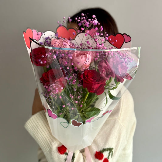 Bouquet of red roses and pink flowers with decorative paper hearts held by a person.