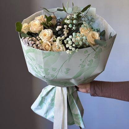 Bouquet of flowers wrapped in green paper with a white ribbon, held against a neutral background.