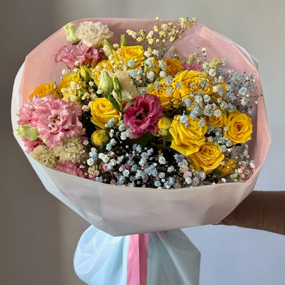 Bouquet of flowers with yellow, pink, and white flowers wrapped in pink paper with a blue ribbon.