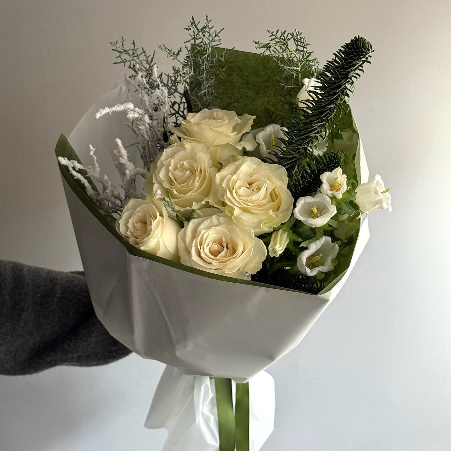 Bouquet of white roses and greenery wrapped in white paper held by a hand against a light background