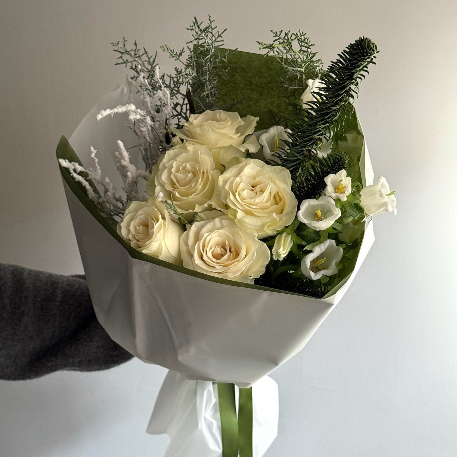 Bouquet of white roses and greenery wrapped in white paper held by a hand against a light background