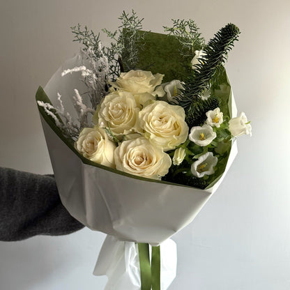 Bouquet of white roses and greenery wrapped in white paper held by a hand against a light background