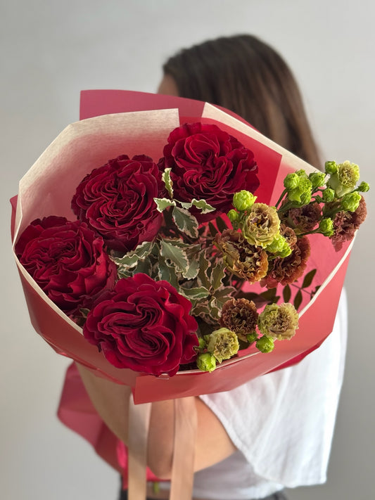 Bouquet of red roses with greenery wrapped in pink paper held by a person.