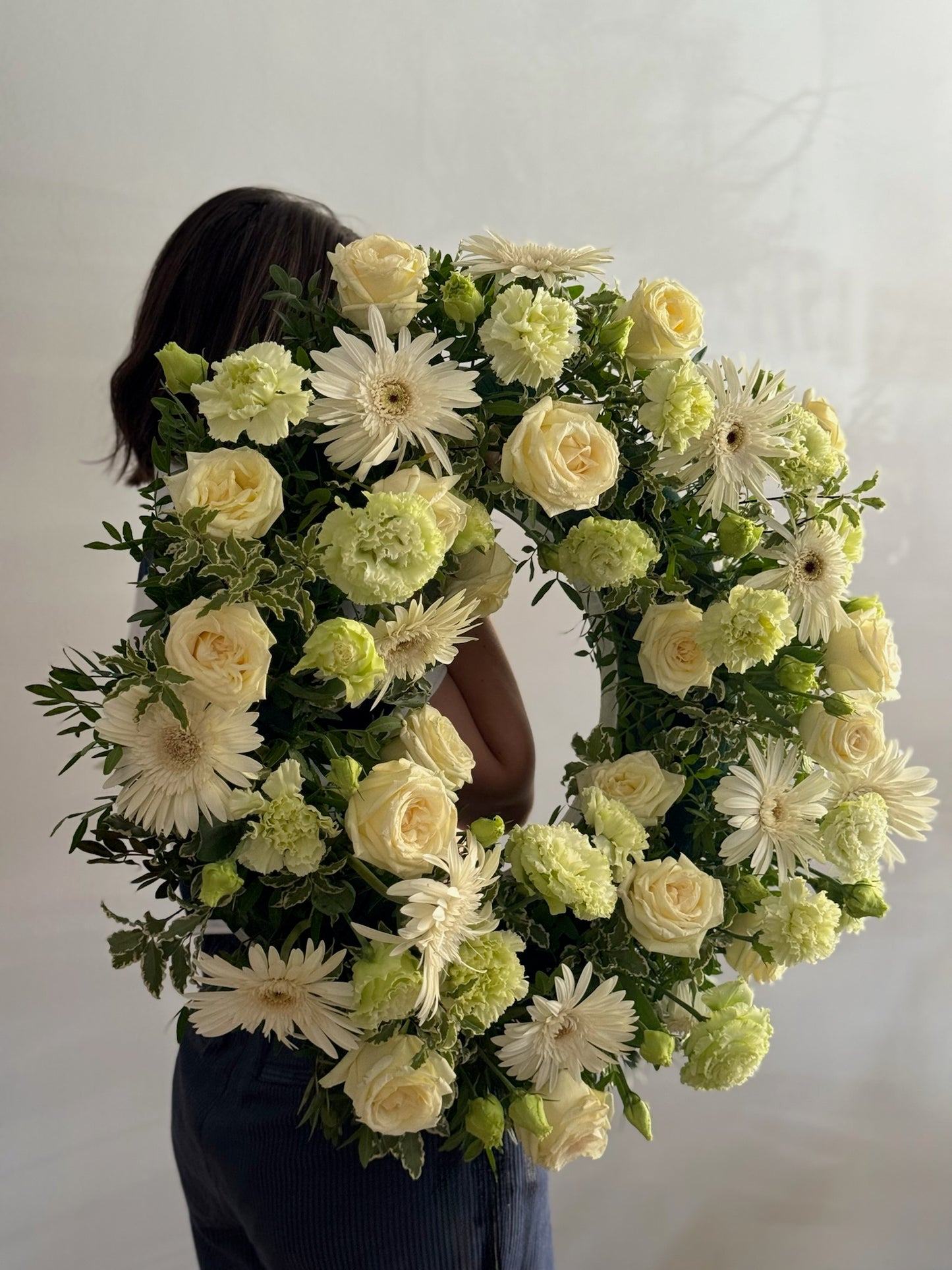 Person holding a large floral wreath made of white and green flowers against a plain background