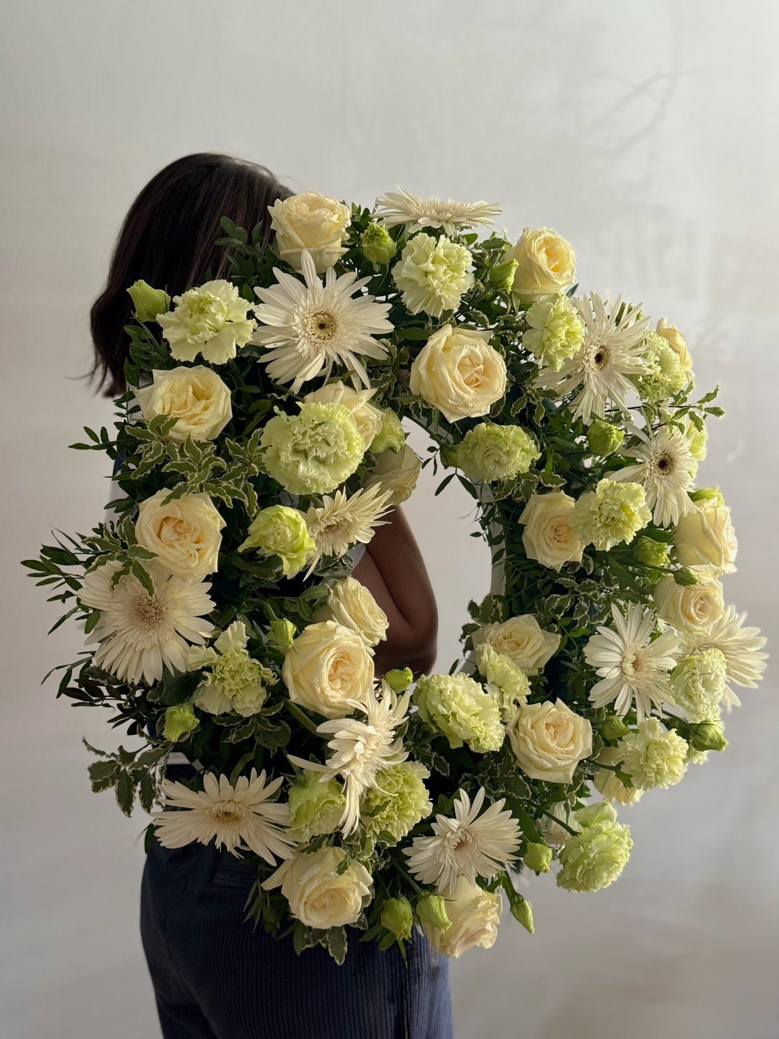 Person holding a large floral wreath made of white and green flowers against a plain background