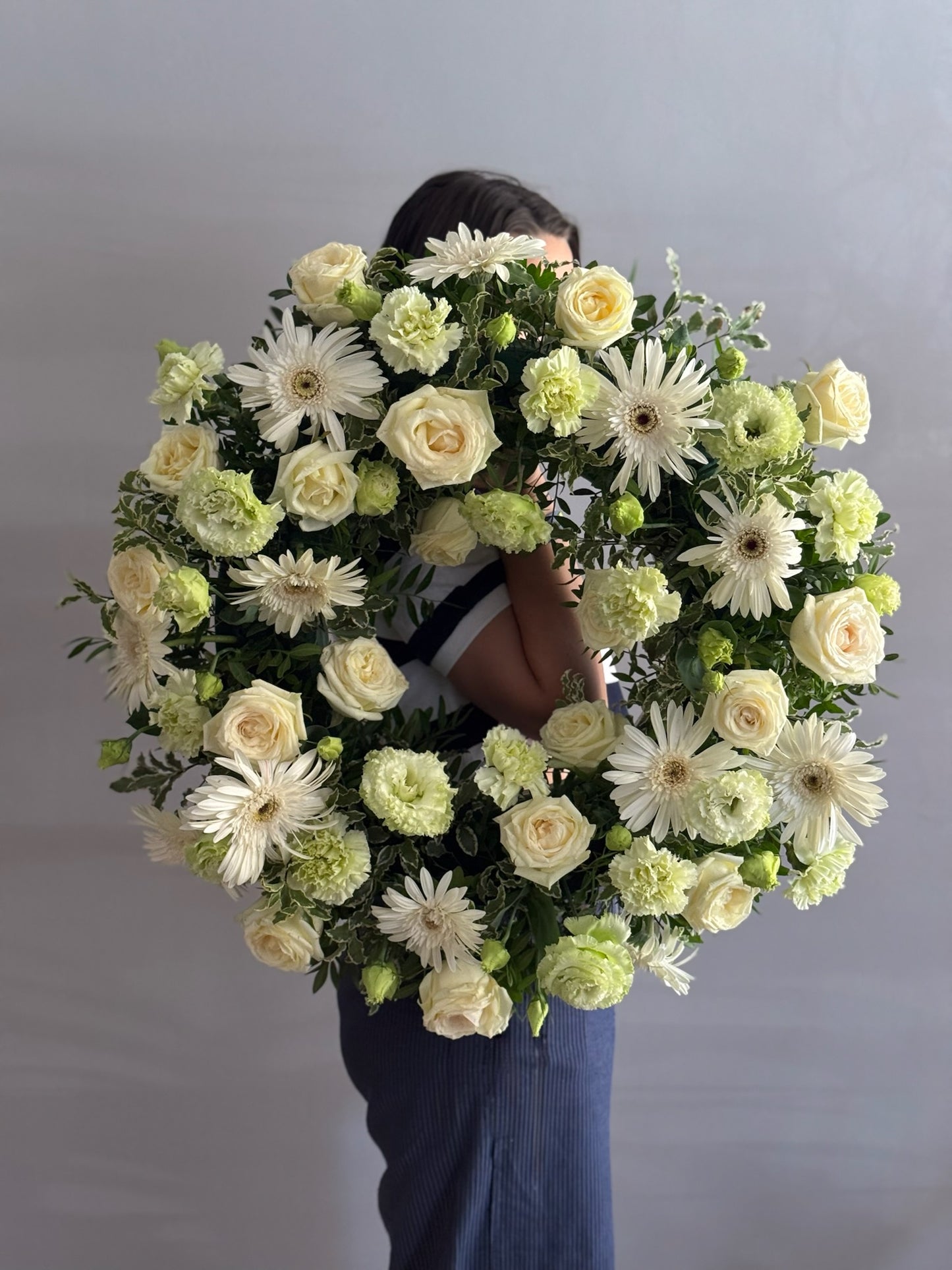 Person holding a large floral wreath with white and green flowers against a plain background