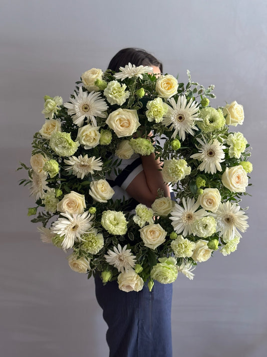 Person holding a large floral wreath with white and green flowers against a plain background