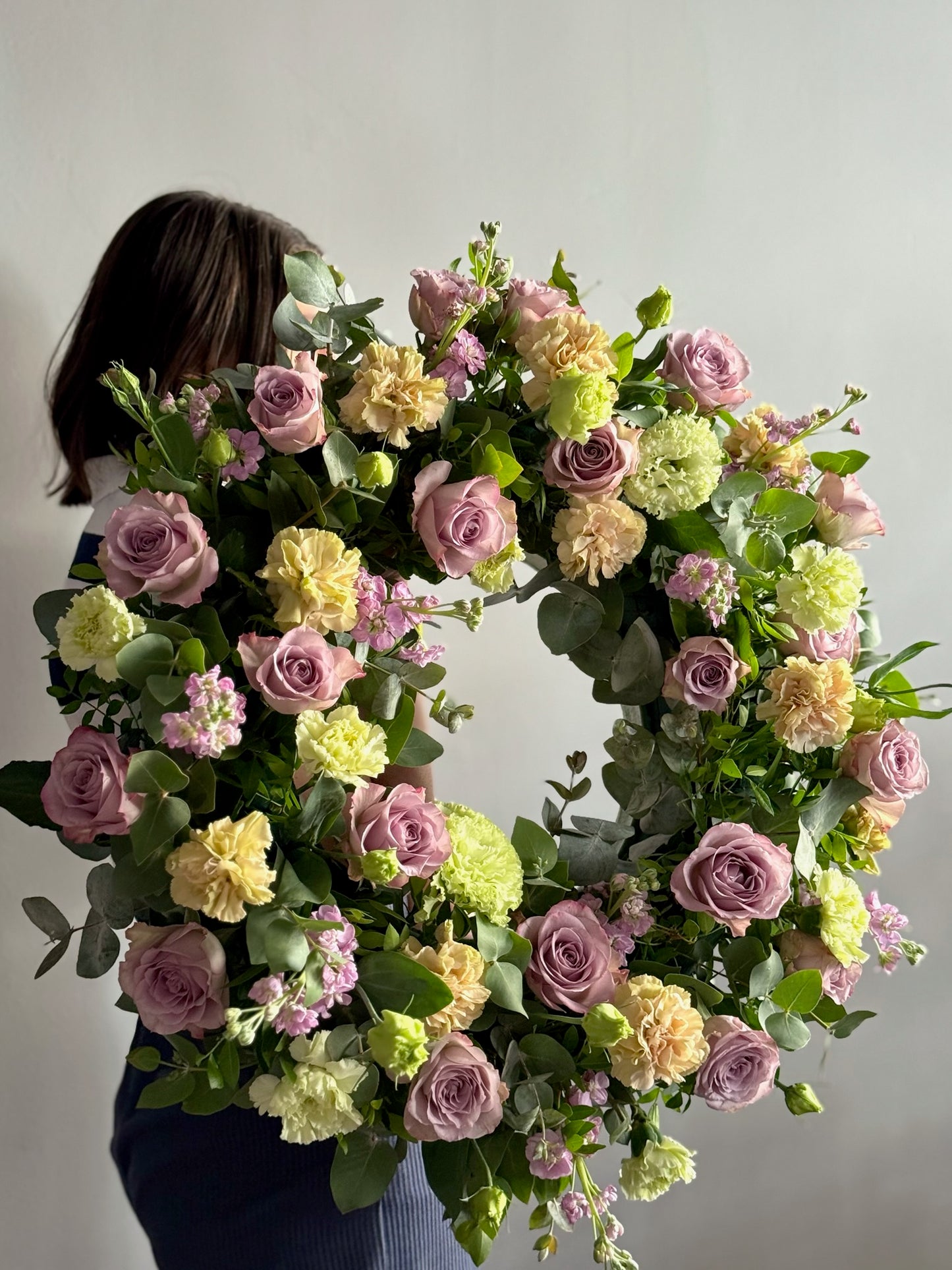 Person holding a large floral wreath with pink, yellow, and green flowers against a plain background