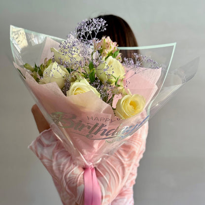 Bouquet of flowers wrapped in pink paper with 'Happy Birthday' text, held by a person.