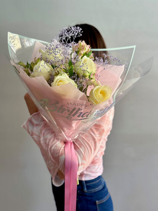 Bouquet of flowers wrapped in pink paper with 'Happy Birthday' text, held by a person.